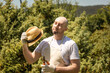 Sunstroke. Portrait of bald bearded man wearing in gloves and apron fanning with straw hat holding a plunger. In the background is a sunny garden trees. Summer heat and gardening