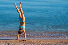 A Child On The Beach Does Sports And Performs Gymnastic Exercises Against The Background Of The Sea. The Girl Does A Handstand At The Water's Edge