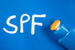 © AliciaFdez - Las letras SPF escritas con protección solar sobre un fondo azul, junto a un bote de protector solar abierto