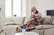 © Studio Romantic - Sick woman who has fever looks frustrated at thermometer which again shows high temperature. Worried woman sitting at home on sofa wrapped in plaid, scarf and wearing hat. Concept of viral infection.