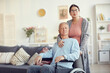© Mediaphotos - Portrait of content loving senior woman with red lips holding handle of wheelchair and embracing handicapped husband in living room