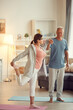 © Mediaphotos - Careful handsome senior husband in tshirt holding hand of wife while assisting her to perform balance pose at home