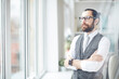 © Mediaphotos - Serious purposeful young bearded businessman in gray waistcoat standing in modern office and looking out window