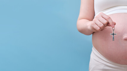 A symbol of the Christian religion in the hands of a pregnant woman, a studio photo on a blue background. Copy space with a place for the text about the law about abortion