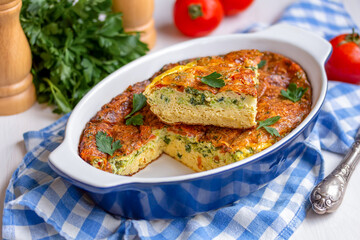 Oven Baked fluffy omelette in a ceramic baking dish on a white wooden table. Sliced vegetable frittata with zucchini or summer squash, cheese, fresh herbs and tomatoes.