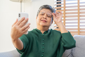 Wall Mural - Presbyopia, Hyperopia mature asian woman holding eyeglasses having problem with vision problem trying to read text on mobile, eye disease of old elderly sitting on couch. Poor eyesight, health care.