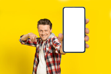 Yo, look at this. Happy handsome man pointing at huge smartphone with white empty screen, wearing red plaid shirt, cellphone display mock up isolated on yellow background. Mobile app advertisement