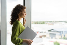 Businesswoman Holding Laptop Looking Out Of Window At Office