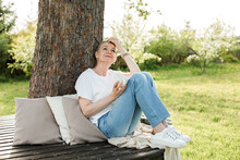 Smiling Woman Holding Coffee Cup Sitting On Bench Around Tree Trunk