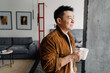 © Drobot Dean - Brunette adult asian man smiling and drinking coffee at home