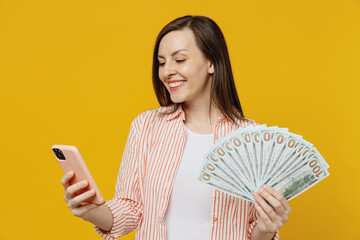 Wall Mural - Young happy woman she 30s in striped shirt white t-shirt holding mobile cell phone fan of cash money in dollar banknotes isolated on plain yellow background studio portrait. People lifestyle concept.