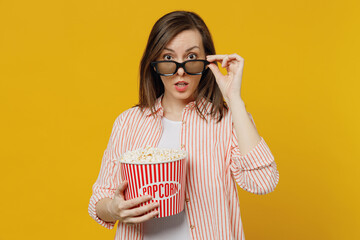 Wall Mural - Young surprised amazed shocked impressed woman lower 3d glasses watch movie film hold bucket of popcorn in cinema point index finger camera on you isolated on plain yellow background studio portrait.