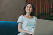 © ViDi Studio - Young happy minded woman she 20s in casual clothes mint t-shirt look aside in reverie mood hold use work on laptop pc computer study online sit on blue sofa indoor rest at home in own room apartment