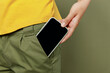 © ViDi Studio - Close up cropped photo shot woman she wear yellow t-shirt holding in hand put into pants pocket mobile cell phone with blank screen workspace area card isolated on plain olive green khaki background
