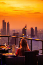 Young Woman With Glass Of Wine Rest At Luxury Rooftop Restaurant Watching Orange Sky Sunset. Female With Cocktail Drink At Sky Bar Terrace Looking At Modern City Skyline. Skyscrapers On Background.