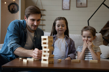 Wall Mural - Joyful father with daughters plays at home. Kids remove wooden blocks from tower. Board game.