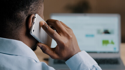 Backside unknown african american man young businessman web designer look at laptop screen surf internet website scroll page interface in social network talk mobile phone wireless call communication
