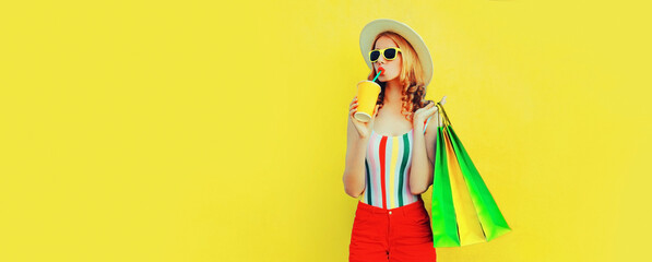 Portrait of happy young woman drinking fresh juice with colorful shopping bags wearing summer straw hat on yellow background