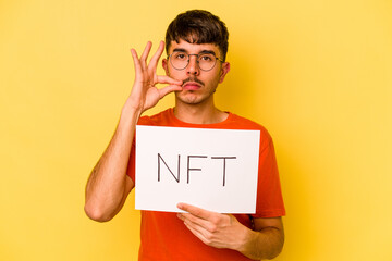 Wall Mural - Young hispanic man holding nft placard isolated on yellow background with fingers on lips keeping a secret.