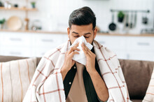 Unhealthy Indian Or Arabian Guy Sits At Home On Sofa In Living Room Under Plaid, Holds Napkin Near Nose, Sneezes, Suffers From Runny Nose, Feels Unwell, Needs Treatment And Doctor Consultation
