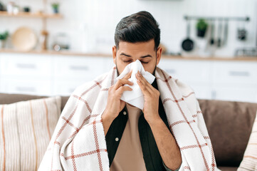 Unhealthy indian or arabian guy sits at home on sofa in living room under plaid, holds napkin near nose, sneezes, suffers from runny nose, feels unwell, needs treatment and doctor consultation