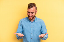 Young Caucasian Man Isolated On Yellow Background Holding Something With Palms, Offering To Camera.