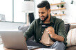 © Kateryna - Indian or Arabian positive young male in casual wear, sitting in cozy living room, holding bank card in hand, using laptop, shopping online, ordering home delivery, paying bills, smiling