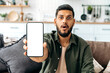© Kateryna - Surprised amazed indian or arabian guy in casual wear, sits on sofa in living room, shows smart phone with empty white mock-up screen, space for advertisement or presentation, looks shocked at camera