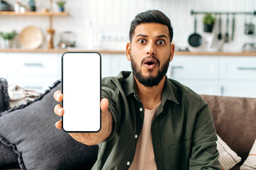 Surprised amazed indian or arabian guy in casual wear, sits on sofa in living room, shows smart phone with empty white mock-up screen, space for advertisement or presentation, looks shocked at camera