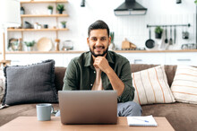 Handsome Confident Attractive Positive Indian Or Arabian Young Man, In Casual Stylish Clothes, Sit At Home In The Living Room On The Sofa, With Laptop, Looks At Camera, Smiling Friendly
