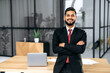 © Kateryna - Positive handsome confident successful Indian or Arabian businessman, in a suit, male entrepreneur, stands in a modern office near the desktop with crossed arms, looks at the camera, smiles friendly