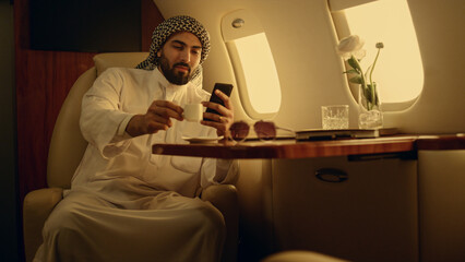 Wall Mural - Happy man looking cellphone in private jet. Smiling arabian rest drinking coffee