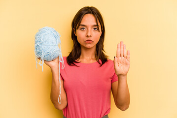 Wall Mural - Young hispanic woman holding a ball of wool isolated on yellow background standing with outstretched hand showing stop sign, preventing you.