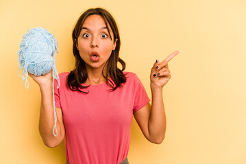 Wall Mural - Young hispanic woman holding a ball of wool isolated on yellow background pointing to the side