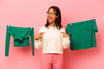 Wall Mural - Young hispanic woman holding laundry basket isolated on pink background person pointing by hand to a shirt copy space, proud and confident