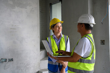 Wall Mural - Young attractive construction man and woman in vest with helmet working with laptop, standing in the under-construction building. Home building project.