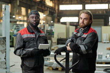 Poster - Small group of young interracial male workers of spacious warehouse in workwear standing in front of camera against workshop interior