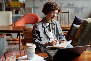 Poster - Young female writer or freelancer with pen and copybook making notes in front of laptop while writing down plan or preparing report