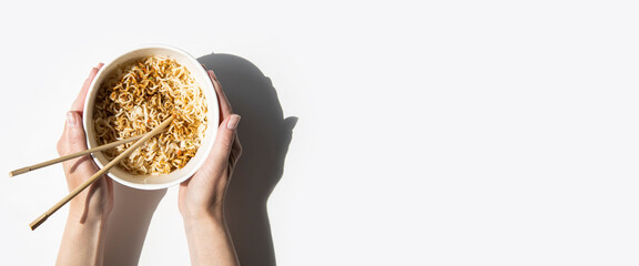 Female hands hold Chinese noodles in a cardboard bowl on a white background. Top view, fly lay. Banner