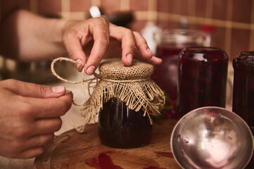 Hands of a housewife, chef confectioner tying bow with a rope around a burlap on a lid of a jar with freshly made cherry berry jam at home kitchen