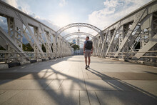Rear View Of Man With Backpack While Walking On Historical Steel Bridge In Singapore Downtown..