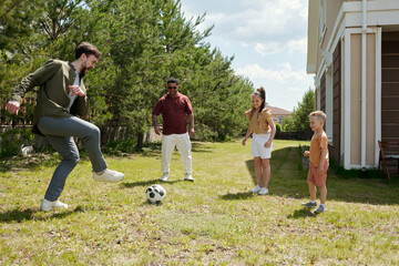 Two young intercultural men and kids playing football on green grass of backyard of their country house on summer day or weekend