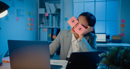 Wall Mural - Young Asian businesswoman sitting on desk overworked tired sleep over a laptop at office at night. Exhausted burnout lady with two post-it over her eyes, adhesive notes on face sleeping at workplace.