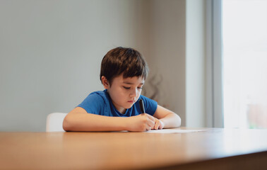 Wall Mural - Kid siting on table doing homework,Child boy holding black pen writing on white paper,Young boy practicing English words at home. Elementary school and home schooling, Distance Education concept