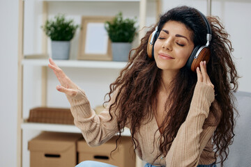 Relaxed calm pleasant tanned curly Latin lady with headphones moves to cool music sit on chair in home modern interior close eyes. Copy space Mockup Banner. Concept meditation. Relax in home