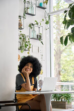 Young Thoughtful Dreamy Smiling African American Student Generation Z Girl With Afro Hair Sitting At Table With Laptop Thinking About Inspiration Ideas In Modern Cafe.
