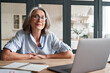 © insta_photos - Smiling stylish mature middle aged woman sits at desk with laptop, portrait. Happy older senior businesswoman, 60s grey-haired lady wearing glasses looking at camera sitting at office table. Headshot.