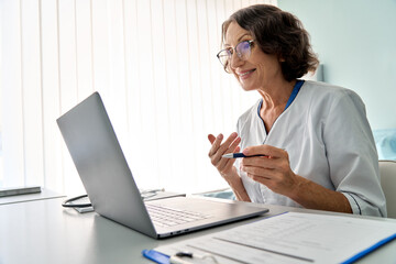 Wall Mural - Senior older female medical worker sitting in modern clinic wearing white doctor's coat having video chat using laptop talking, consulting remotely. Telemedicine distant healthcare concept.