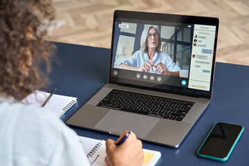 Girl student studying virtual course with remote teacher or tutor, learning distance class training, looking at laptop computer watching online webinar sitting at home table. Over shoulder screen view