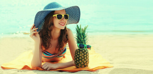 Summer portrait of happy smiling young woman lying on the beach with funny pineapple wearing straw hat